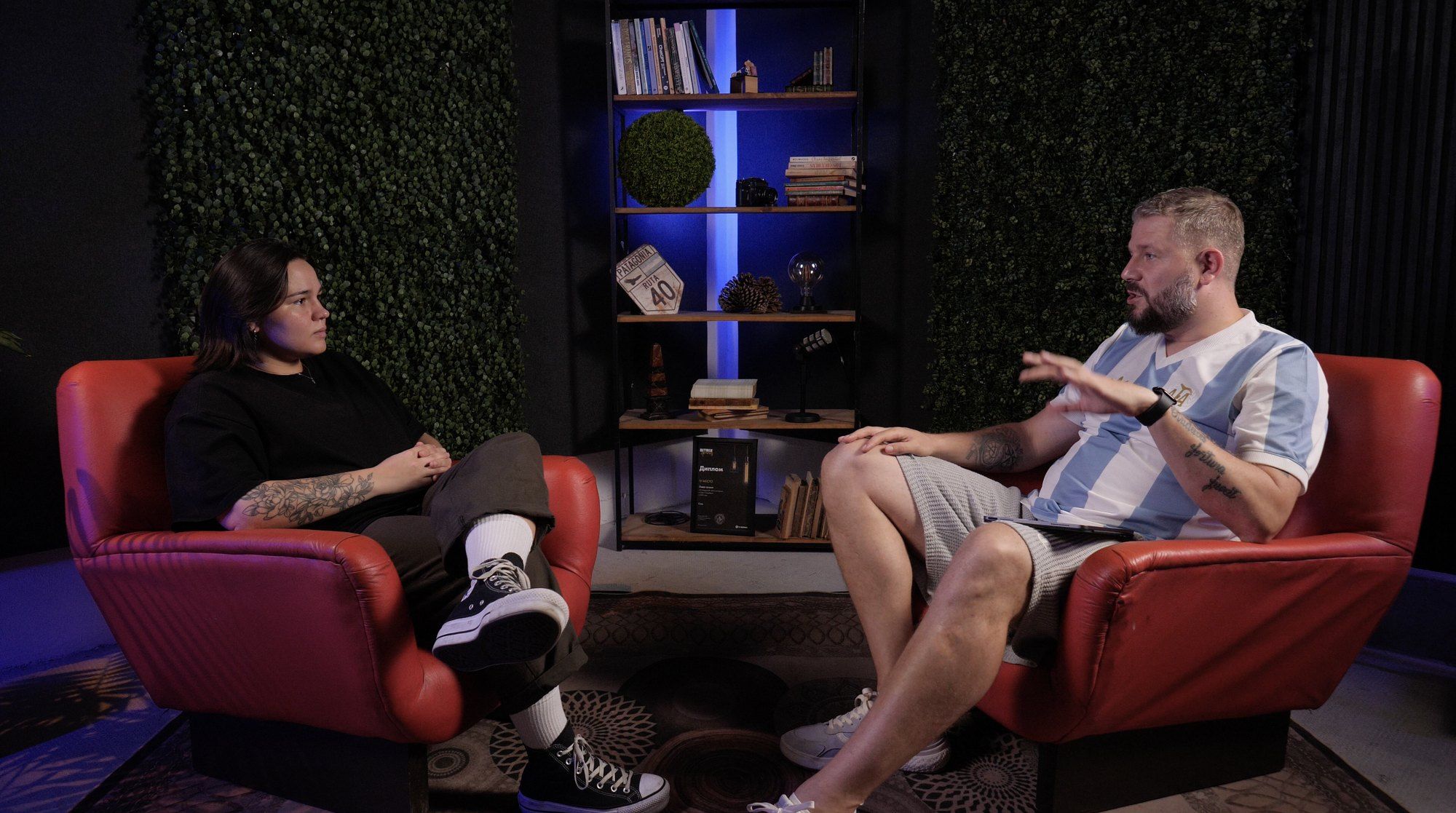 Toma de podcast en el estudio Liberty: dos participantes en sillones, paredes verdes y una estantería iluminada entre ellos